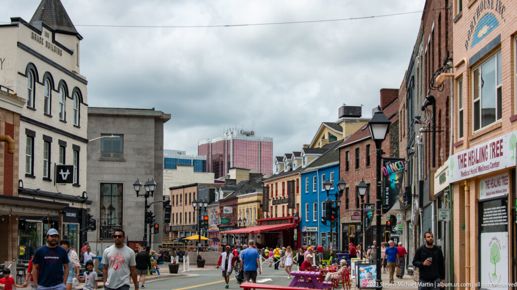 Busy downtown St. John’s street lined with colorful historic buildings, shops, pedestrians, patios, and a distant office tower.