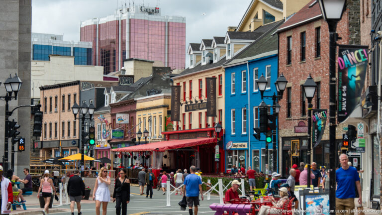 Pedestrians walk past blue, red, yellow, and brick storefronts on a lively downtown St. John’s street.