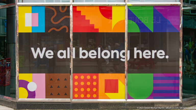 Storefront window with colorful geometric graphics and large white text reading “We all belong here.”