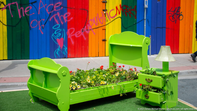 Bright green bed planted with flowers, armchair, nightstand, and lamp before a rainbow-painted wall.