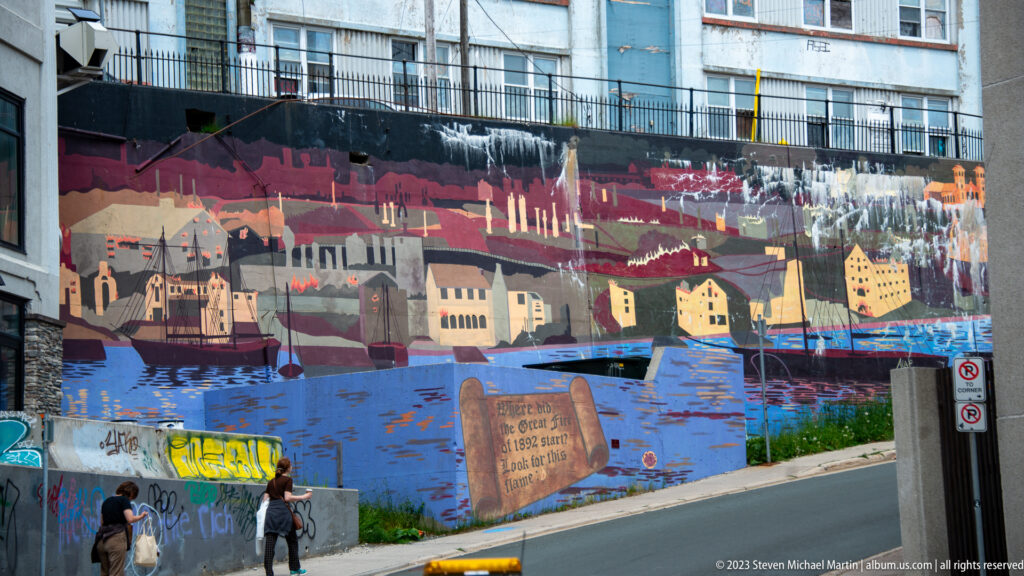 Large mural of historic St. John’s waterfront buildings and ships painted across a retaining wall above pedestrians.