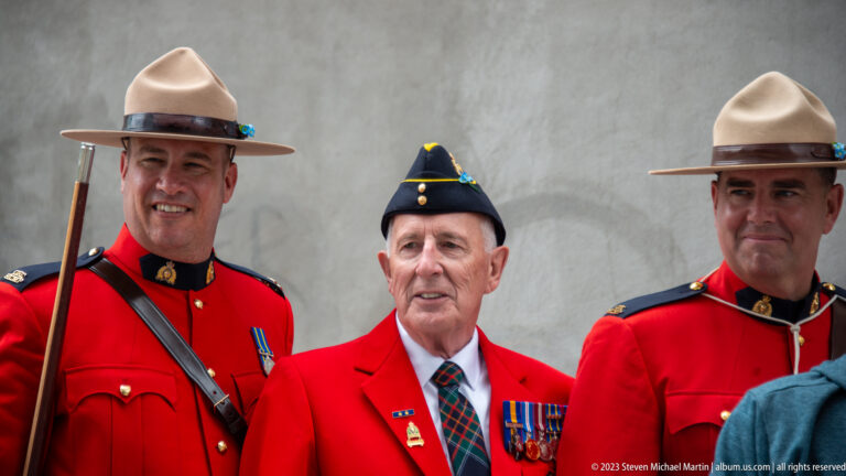 Two RCMP officers in red serge pose with an older veteran wearing a red blazer, service cap, and medals.