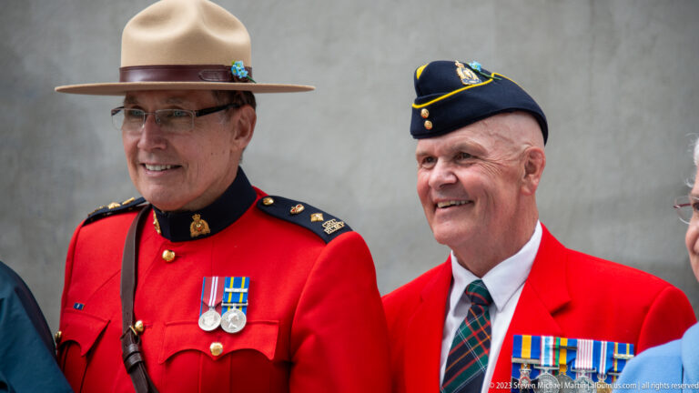 Smiling RCMP officer in red serge beside a decorated veteran in a red blazer and service cap.