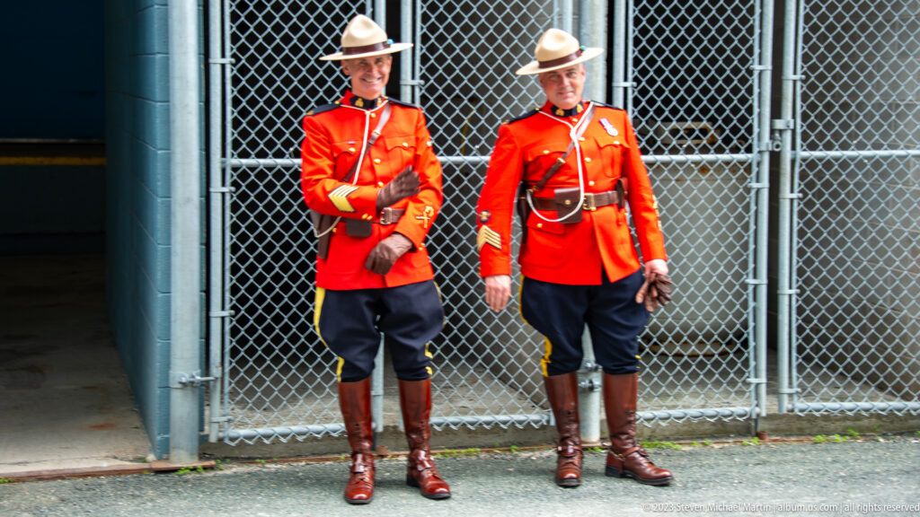 Two RCMP officers stand before a chain-link gate in red serge, dark breeches, campaign hats, and tall boots.