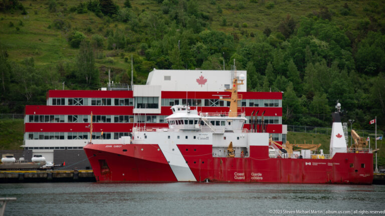 Red-and-white Canadian Coast Guard vessel moored beside a red-and-white waterfront facility beneath a wooded hillside.