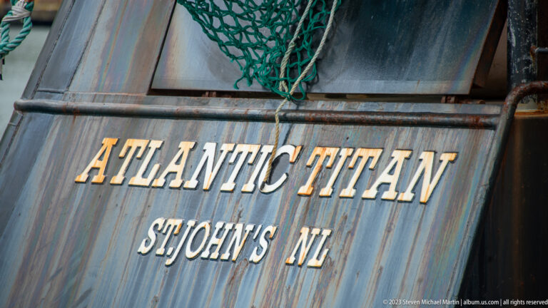 Gold lettering reading “Atlantic Titan, St. John’s NL” on a weathered dark vessel stern beneath green netting.