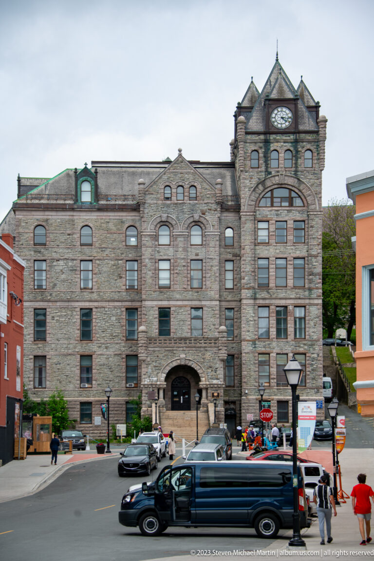 Large gray-stone historic building with arched entrance, many windows, and a clock tower at a downtown intersection.
