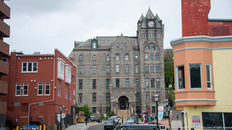 Stone clock-tower building seen at the end of a narrow downtown St. John’s street between colorful façades.