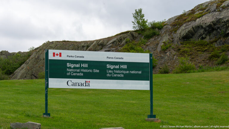 Bilingual Parks Canada sign for Signal Hill National Historic Site standing on grass below a rocky hillside.