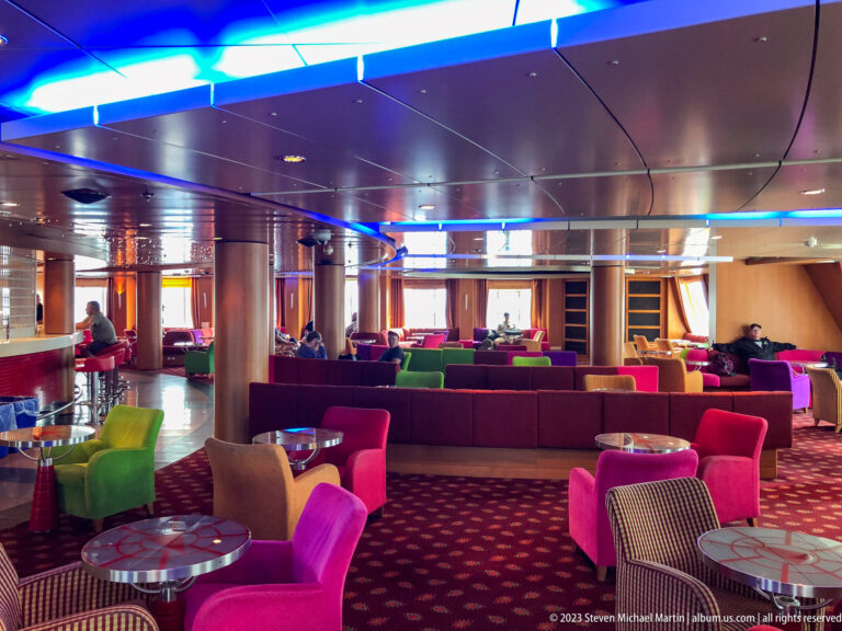 Ferry passenger lounge with brightly colored armchairs, small tables, patterned carpet, windows, and blue ceiling lighting.