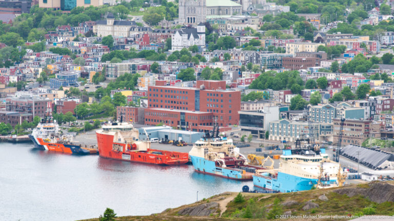 Red, orange, and blue offshore vessels moored along the St. John’s waterfront below downtown buildings.