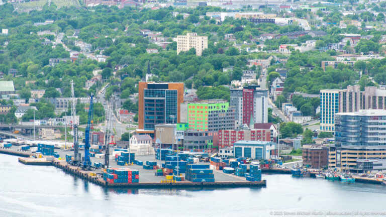 Container stacks, cranes, service vessels, and modern buildings along the St. John’s waterfront below a green hillside.