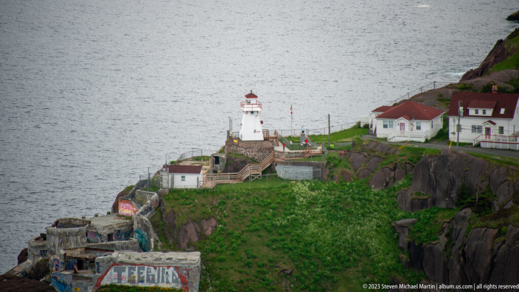 Telephoto view of Fort Amherst lighthouse, red-roofed buildings, stairs, green slope, and gray Atlantic water.