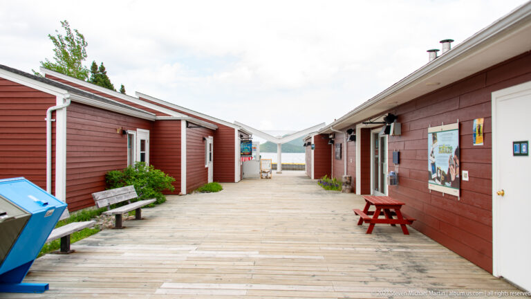 Wooden boardwalk corridor between red visitor-centre buildings with benches, posters, and picnic tables.
