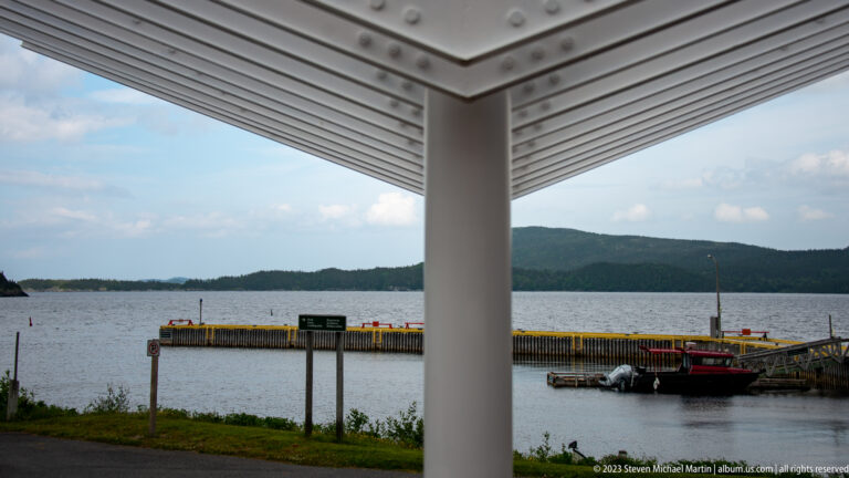 View from a white canopy toward a dock, red boat, sheltered water, and forested hills.