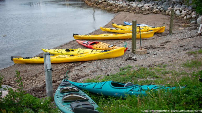 Yellow, orange, and turquoise sea kayaks lined along a gravel shore beside calm water.