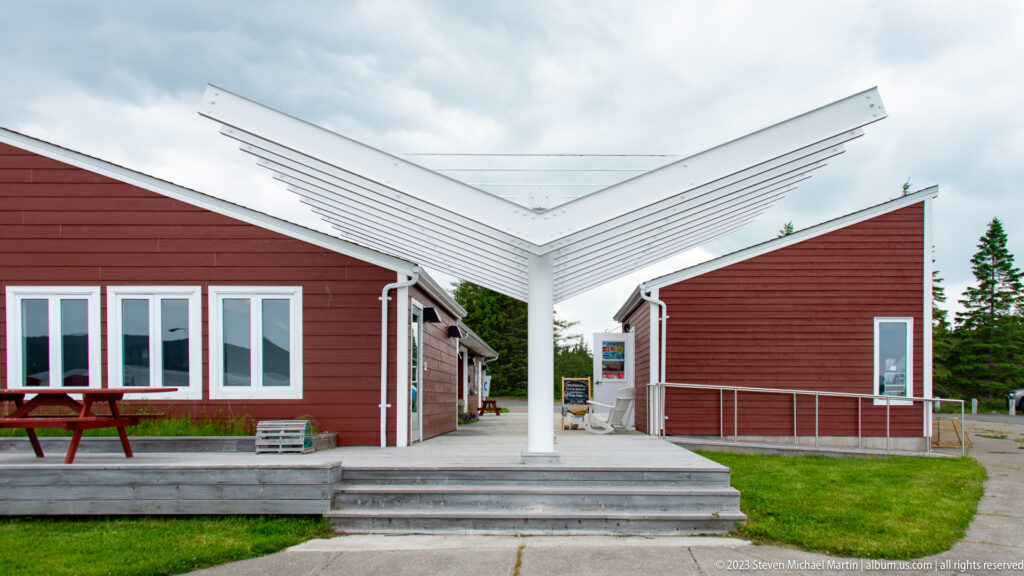 Angular white entrance canopy between red visitor-centre buildings with steps, ramp, and picnic table.