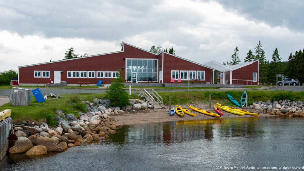 Red visitor centre behind a rocky inlet, with yellow kayaks gathered at the shoreline.