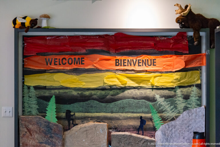 Bilingual Welcome Bienvenue display with colorful fabric, forest scenery, cutout trees, rocks, people, and a moose figure.