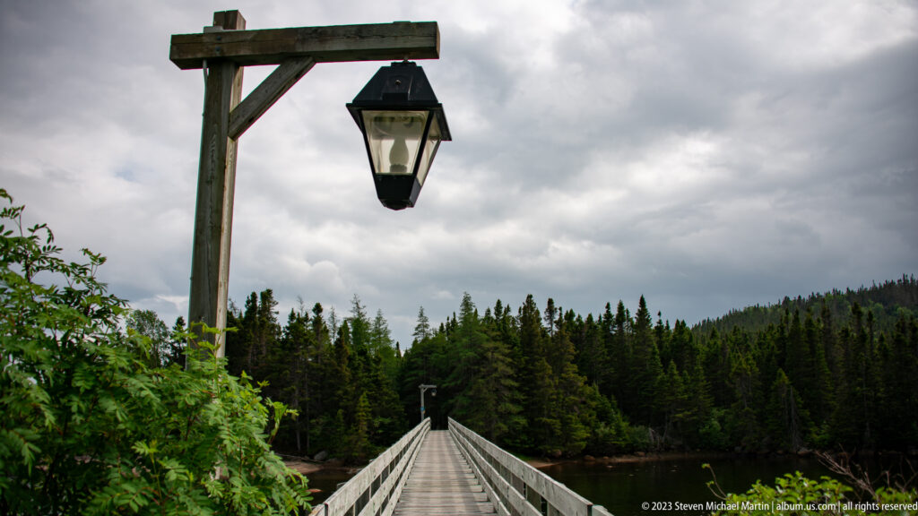 Long wooden footbridge with railings and a hanging lantern crossing sheltered water toward dense evergreen forest.