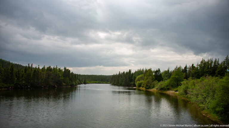 Calm narrow waterway bordered by dense forest beneath dark storm clouds and a brighter distant sky.
