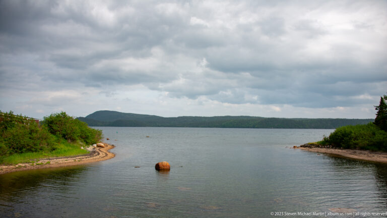 Sheltered bay with rocky low shores, a rust-colored buoy, forested ridge, and cloudy sky.