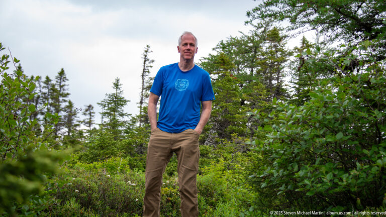 Adult man in a blue T-shirt and tan trousers standing on a narrow path among dense evergreen and leafy vegetation.
