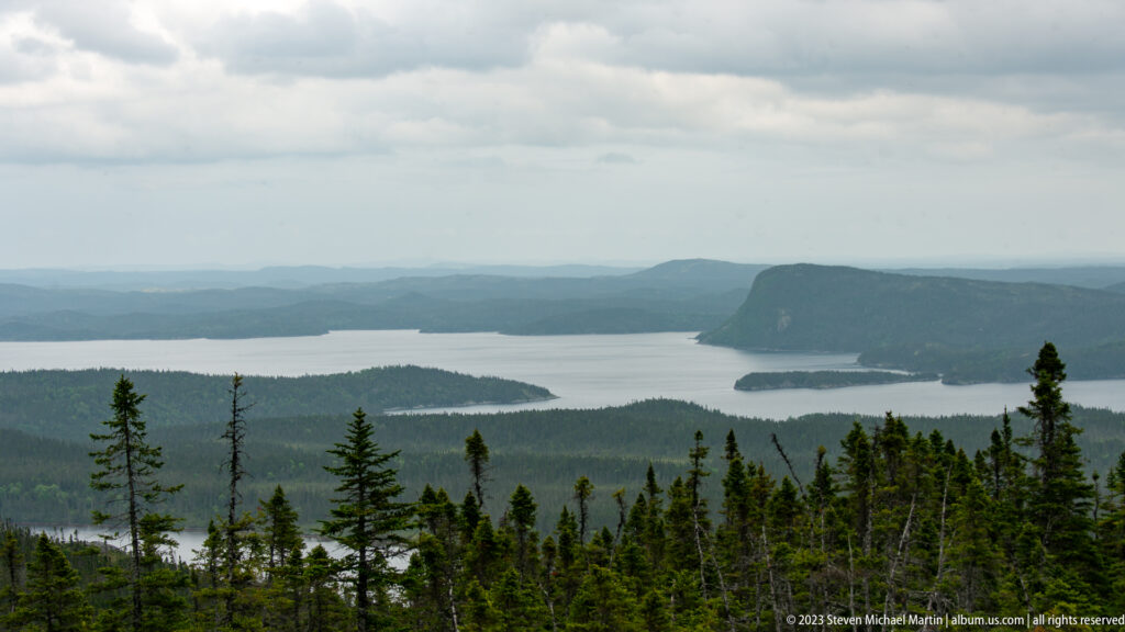Telephoto coastal view with forested headland, low islands, pale inlet water, and hazy ridges beyond.
