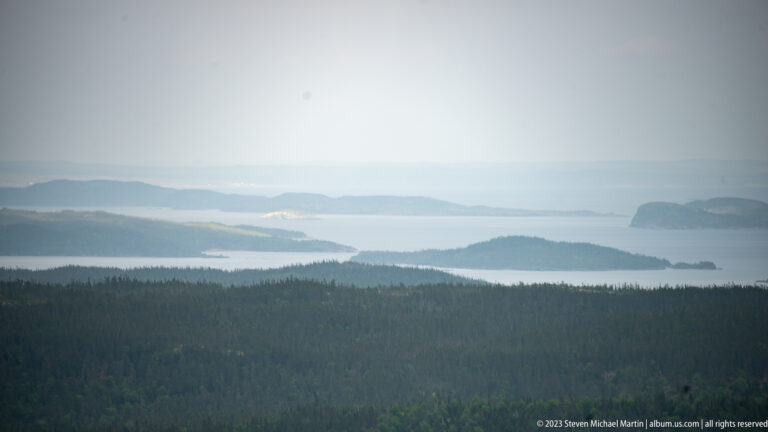 Hazy long-distance view over dark forest to pale water with several low islands and distant shoreline.