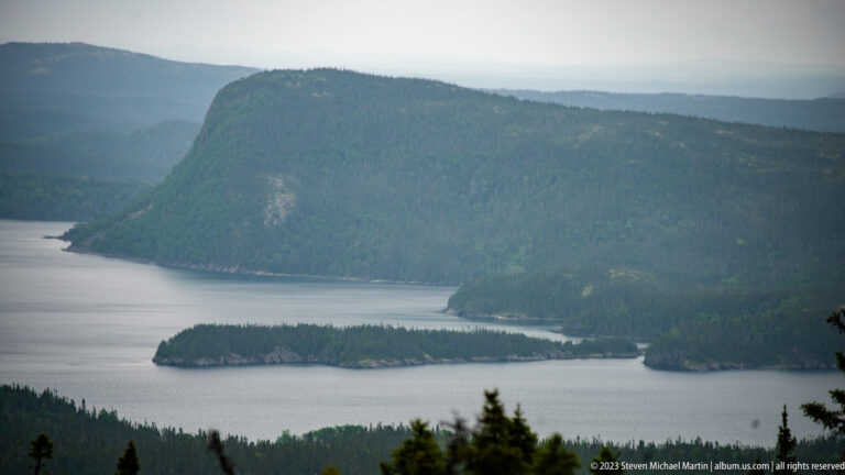 Narrow forested island in calm water beneath a steep wooded shoreline with patches of exposed rock.