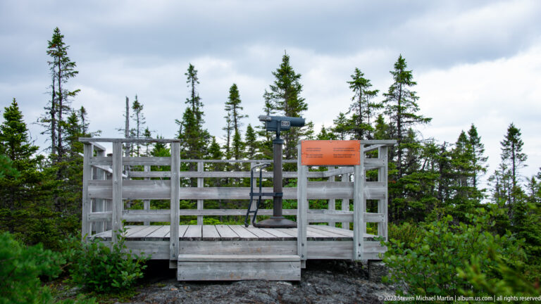 Small wooden viewing platform on rocky ground among conifers, with railings, an orange sign, and a dark post.