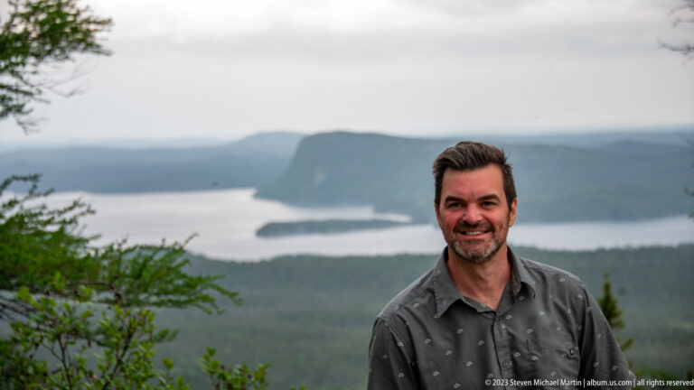 Adult man at an elevated overlook with blurred forest, islands, water, and a broad headland behind him.