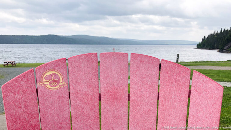 Bright red Parks Canada Adirondack chair facing calm water, a picnic table, forested shore, and low hills.