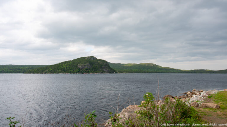 Rocky shoreline across open water from a rounded forested headland beneath heavy gray clouds.