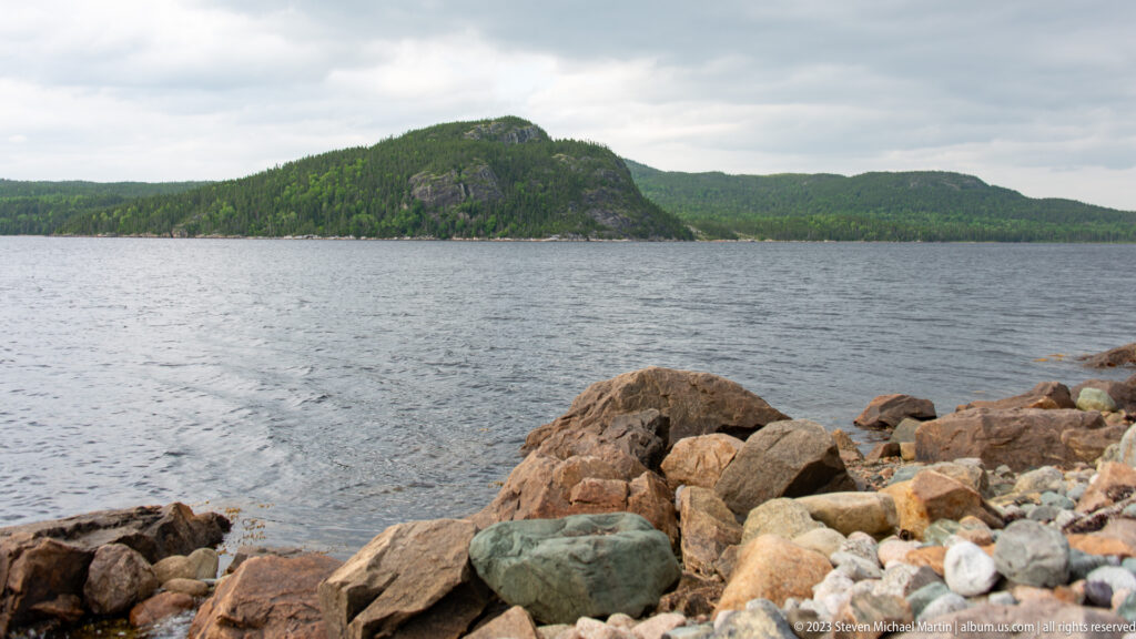 Large shoreline rocks and pebbles beside rippled water, with a steep forested headland across the bay.