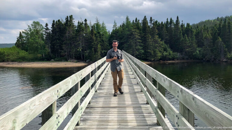 Adult man walking toward the camera on a wooden footbridge over calm water, with dense forest behind.