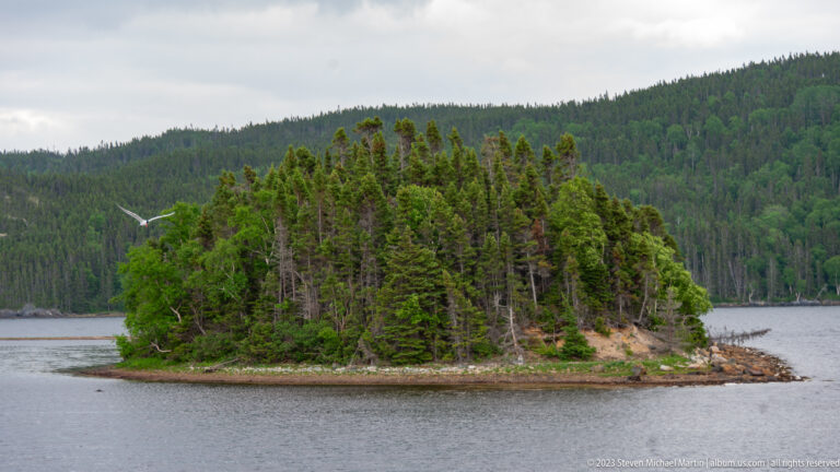 Small densely forested island with a narrow rocky shore in calm water before a larger wooded hillside.