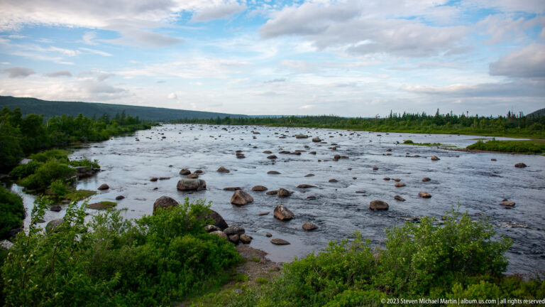 Wide shallow river filled with rounded boulders, bordered by low green vegetation beneath a partly cloudy sky.