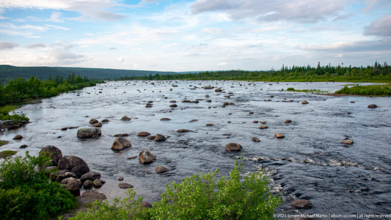 Elevated view over a broad rocky river with many exposed boulders, green banks, and broken clouds.
