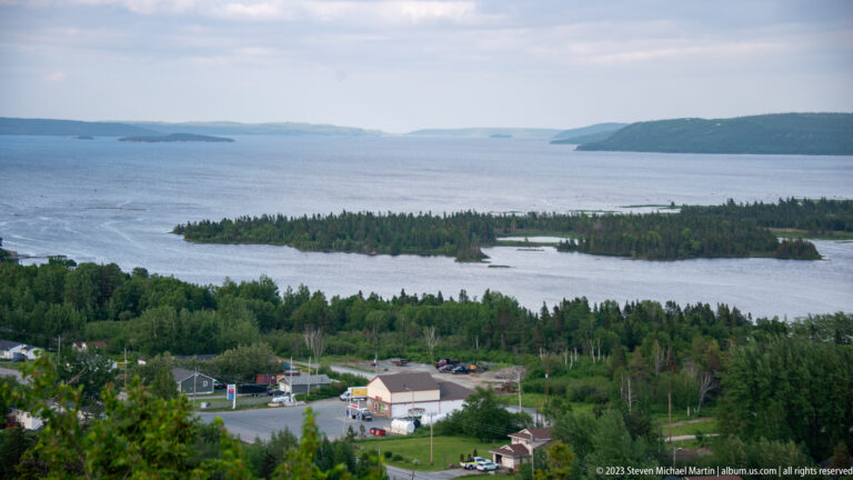 Elevated view over a small settlement and forest toward wooded islands, sheltered water, and distant headlands.