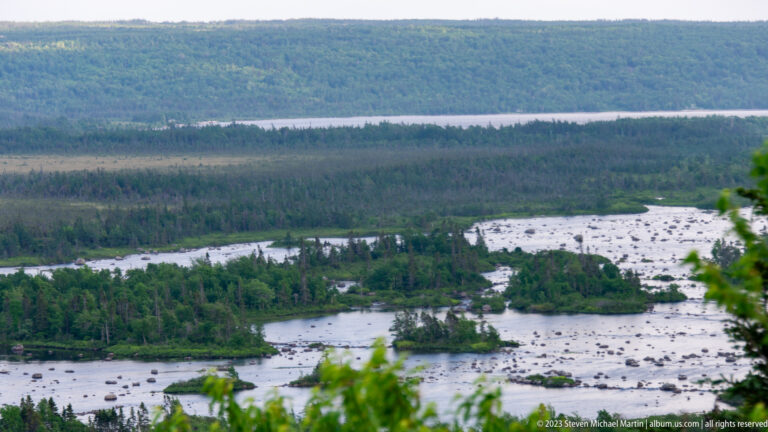 Elevated view of a broad river divided by wooded islands and many exposed boulders, with forested ridge beyond.