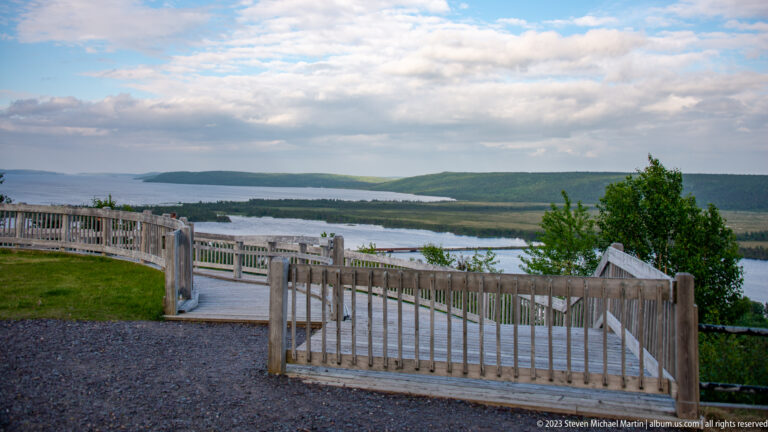 Wooden accessible viewing platform with railings overlooking water, islands, low hills, and a cloudy sky.