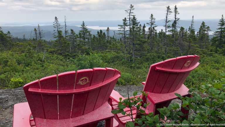 Two red Parks Canada Adirondack chairs facing a forested overlook with distant water and low hills.