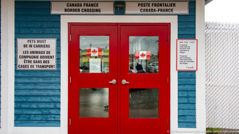 Red doors with Canadian flags and bilingual border-crossing signs on a blue wooden frontier building.