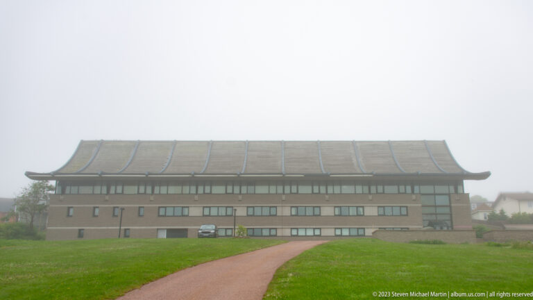 Large symmetrical building with a wide upturned roof, long windows, lawn and central path in fog.