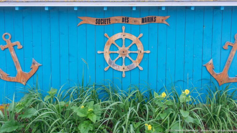 Blue wooden wall decorated with anchors, a ship’s wheel and a sign reading SOCIETE DES MARINS above green plants.