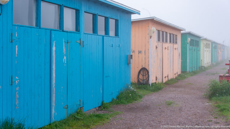 Row of blue, salmon, green and pale wooden sheds lining a gravel lane in fog.