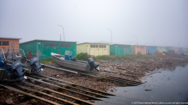 Open motorboats with outboard engines sit on wooden launching rails beside colorful sheds on a foggy rocky shore.
