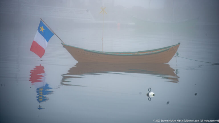 Small brown wooden boat with green trim and an Acadian flag floating on calm foggy water.