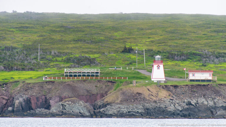 Small white-and-red navigation light with service building and walkway on a rocky green shoreline.